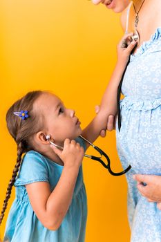 Motherhood, hot summer - croped portrait pregnant mother unrecognizable woman blue dress little Daughter girl sibling treat mom role play game stethoscope unborn big belly tummy on yellow background.