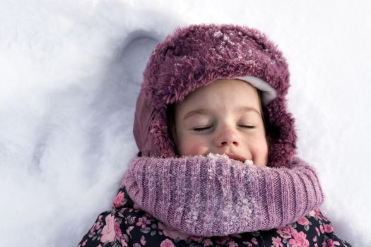 Winter, family, childhood concepts - close-up portrait authentic little preschool minor girl in pink clothes smile laugh closing eyes laying on snow in frosty weather day outdoors. Funny kid face.