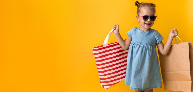 Portrait Beautiful Happy Little Preschool Girl In Sunglasses Smiling Cheerful Holding Cardboard Bags Isolated On Orange Yellow Studio background. Happiness, Consumerism, Sale People shopping Concept.