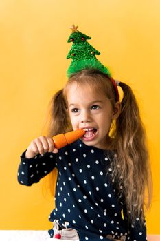 Portrait positive cheerful smiling happy little schoolgirl girl Christmas tree decoration polka dot dress biting eat orange carrots on orange background. New year, holiday, celebration, winter concept.