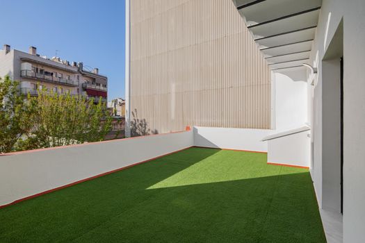 Empty sunny terrace of refurbished apartment with artificial green grass lawn. View to the street and blue sky in the center of Barcelona, Spain