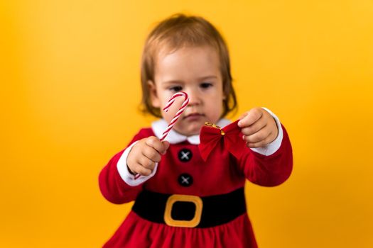 Blured Portraite Cute Happy Baby Girl in Santa Suit Holding Butterfly Caramel Candy At Yellow Background. Child Play Christmas Scene Celebrating Birthday. Kid Have Fun Spend New Year Time Copy Space.