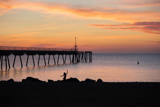 Silhouette of a woman dancing at the beach in the morning. Sunrise in the mediterranean sea with view to the pontoon. Pont del Petroli, Badalona, Barcelona, Catalonia, Spain.