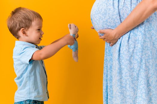 Motherhood, love, Childhood, hot summer - croped portrait pregnant unrecognizable mother woman blue dress little boy son sibling show teddy bear toy big belly brother on yellow background copy space.