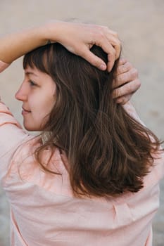 A woman in pink shirt holding her head with natural hair looking away. Brunette girl with long hair, view from the back