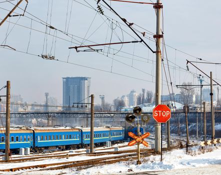A red stop sign at a railroad crossing against the backdrop of railroad tracks, blue train carriages and cityscape at the entrance to a winter city.