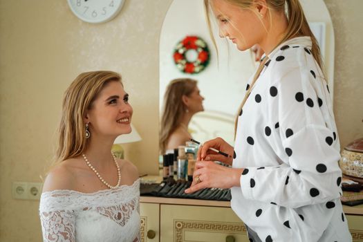 Make-up artist makes professional makeup for a young woman, a bride. A smiling bride in a white dress, pearl necklace and earrings looks at the makeup artist
