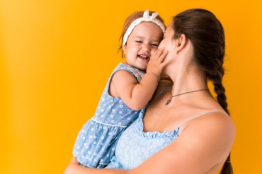 Motherhood, hot summer - croped portrait young pregnant happy smiling mother woman in blue dress holding his little baby daughter toddler sibling in arms looking into eyes kisses on yellow background.