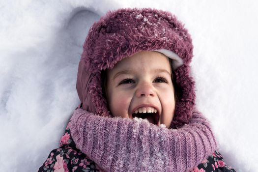 Winter, family, childhood concepts - close-up portrait authentic little preschool girl in pink clothes smile laugh shout with open mouth laying on snow in frosty weather day outdoors. Funny kid face.