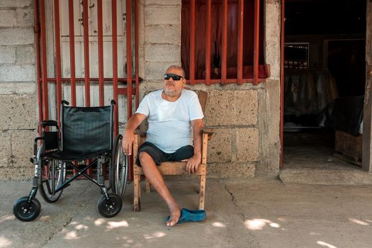 Disabled mature Latin man without a leg resting outside his house with dark glasses and his wheelchair next to him.
