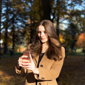 Beautiful teen girl in the Park, holding smartphone and chatting online on Internet, takes a selfie. Young woman with long hair.