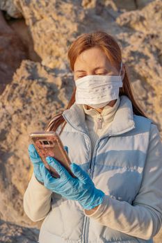 Red-haired girl in a mask looks at the phone, dressed in bright clothes on the street on a sunny day, protective pandemic