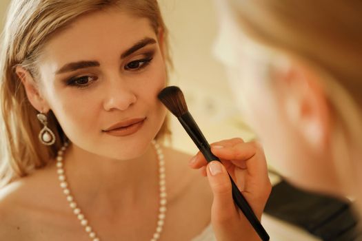 Make-up artist makes professional makeup of a young woman, a bride. The bride in a white dress, a pearl necklace and earrings. Make-up artist applies powder