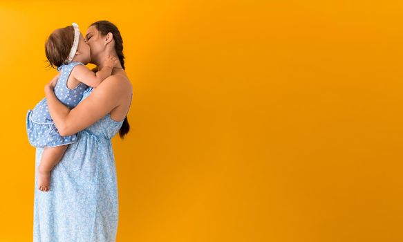 Motherhood, hot summer - Banner young pregnant happy smiling mother woman in blue dress holding little baby daughter toddler sibling in arms looking into eyes kisses on yellow background coppy space.