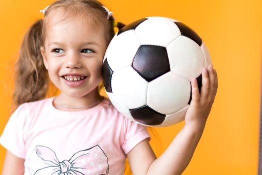 Authentic cute smiling preschool little girl with classic black and white soccer ball look at camera on yellow background. child play football in t-shirt and shorts. Sport, championship, team concept.