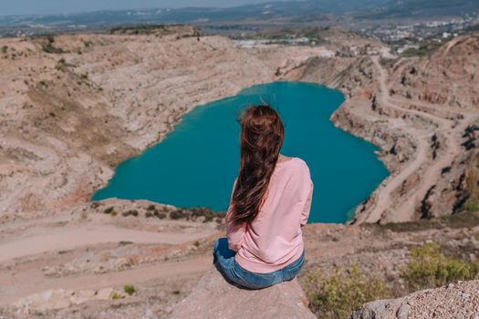 A red-haired girl with loose hair in a pink sweatshirt and jeans sits against the background of an azure lake in the form of a heart, rear view.