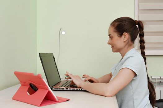 A young beautiful woman uses a laptop while sitting at her workplace, a cheerful smiling girl. Technology, business, homework, online learning, concept of learning, communication with loved ones quarantine distance education