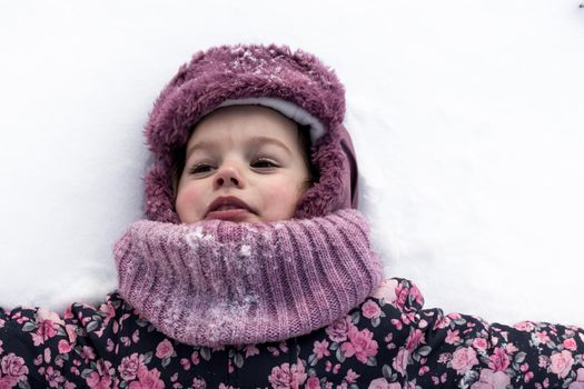 Winter, family, childhood concepts - close-up portrait authentic little preschool minor girl in pink clothes smile laugh closing eyes laying on snow in frosty weather day outdoors. Funny kid face.
