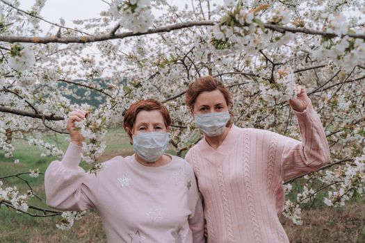 Mom and daughter in medical masks in pink sweaters are standing in a flowering cherry garden. Quarantine concept, next to the house.