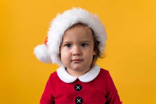 Smiling Portraite Cute Happy Cheerful Chubby Baby Girl in Santa Hat Looking On Camera At Yellow Background. Child Play Christmas Scene Celebrating Birthday. Kid Have Fun Spend New Year Time Copy Space.