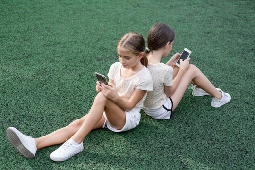 two pretty girls in identical clothes using phone on sports ground or athletic field. kids are surfing in internet. smartphone addiction. advanced, modern children in social networks.