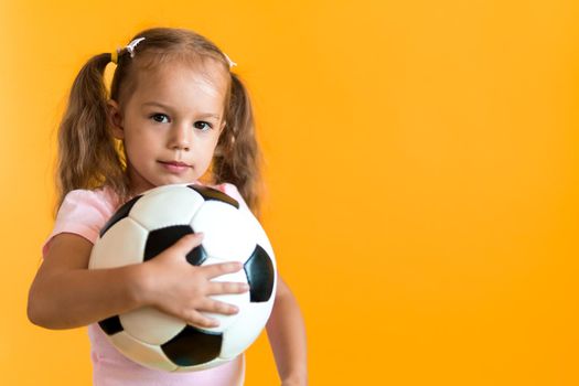 Authentic cute smiling preschool little girl with classic black and white soccer ball look at camera on yellow background. child play football in t-shirt and shorts. Sport, championship, team concept.