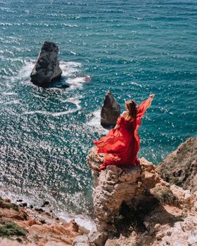 A girl with loose hair in a red dress waved her skirt on the yellow rocks overlooking the sea. Sunny path on the sea from the rising sun