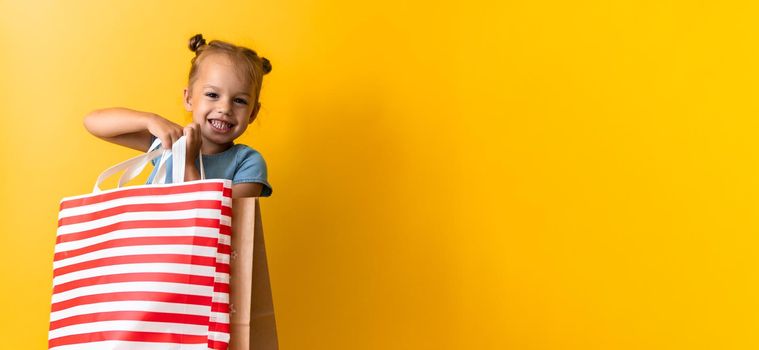 Banner Portrait Caucasian Beautiful Happy Little Preschool Girl Smiling Cheerful And Holding Cardboard Bags Isolated On Orange Yellow Background. Happiness, Consumerism, Sale People shopping Concept.