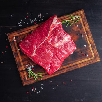 Big whole piece of raw beef meat with seasoning on chopping board on dark background with rosemary, seasonings, top view.