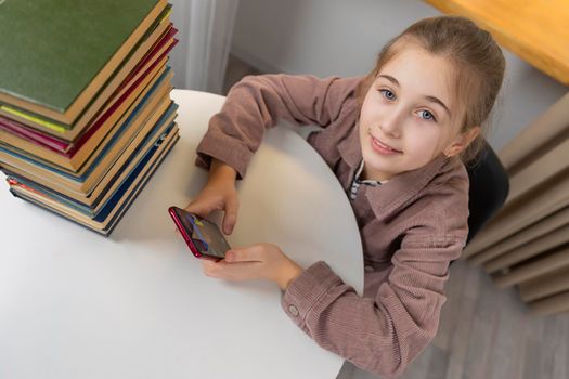 girl sits at a table on which there are books and looks at the phone