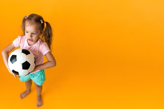 Authentic cute smiling preschool little girl with classic black and white soccer ball look at camera on yellow background. child play football in t-shirt and shorts. Sport, championship, team concept.