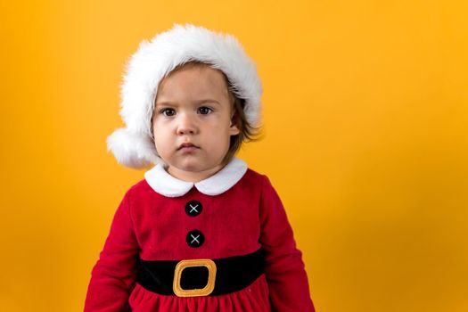 Serious Portraite Cute Happy Cheerful Chubby Baby Girl in Santa Hat Looking On Camera At Yellow Background. Child Play Christmas Scene Celebrating Birthday. Kid Have Fun Spend New Year Time Copy Space.