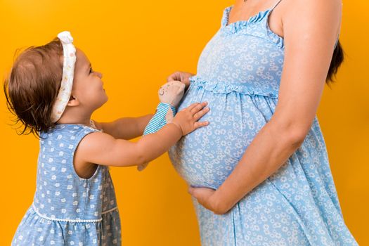 Motherhood, love, Childhood, hot summer - croped portrait pregnant mother unrecognizable woman blue dress little sibling girl sister hug touch mum big belly brother on yellow background copy space.