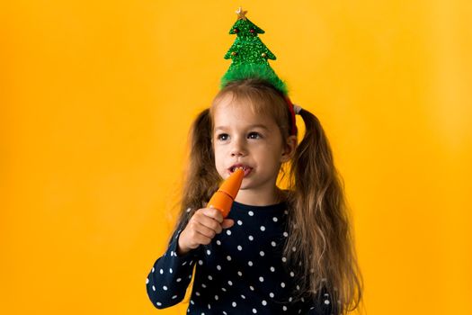 Portrait positive cheerful smiling happy little schoolgirl girl Christmas tree decoration polka dot dress biting eat orange carrots on orange background. New year, holiday, celebration, winter concept.