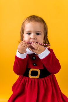Portraite Cute Cheerful Chubby Baby Girl in Santa Suite Holding Eating Caramel Candy At Yellow Background. Child Play Christmas Scene Celebrating Birthday. Kid Have Fun Spend New Year Time Copy Space.