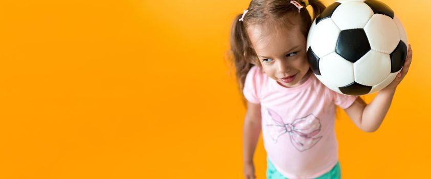 Authentic cute smiling preschool little girl with classic black and white soccer ball look at camera on yellow background. child play football in t-shirt and shorts. Sport, championship, team concept.