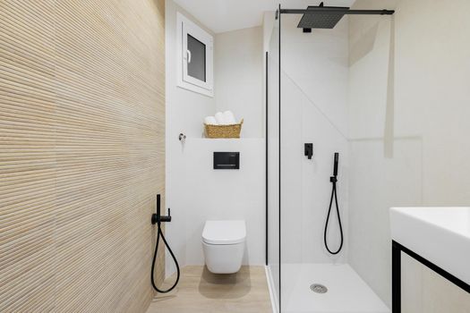 Interior of modern style bathroom in white and beige colors in refurbished apartment. Shower zone and toilet, with black faucets, towels and tiled floor and walls