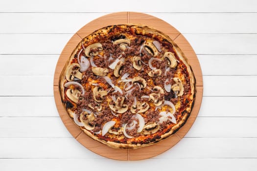 Whole round pizza on wooden plate topped with mushrooms, onion and chopped meat. Top view on white board background with copy space.