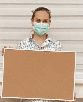 A young woman in a mask holds an empty cork board in her hands, standing on a white background. Copy space on whiteboard for image or message. Young woman. Concept of paying attention to your promise