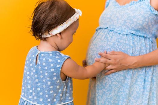 Motherhood, love, Childhood, hot summer - croped portrait pregnant mother unrecognizable woman blue dress little sibling girl sister hug touch mum big belly brother on yellow background copy space.