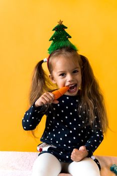 Portrait positive cheerful smiling happy little schoolgirl girl Christmas tree decoration polka dot dress biting eat orange carrots on orange background. New year, holiday, celebration, winter concept.