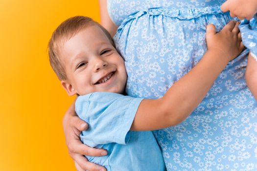 Motherhood, femininity, love, Childhood, hot summer - croped portrait pregnant unrecognizable woman blue dress little preschool boy son sibling hug mum big belly brother on yellow background copy space.