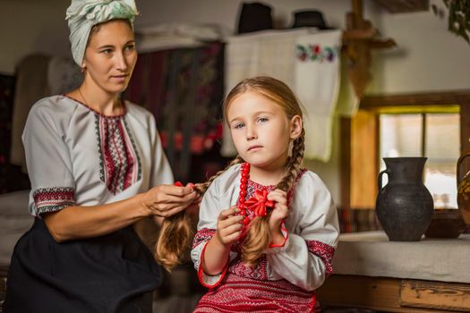 mother combing her daughter's hair, both dressed in Ukrainian national costumes