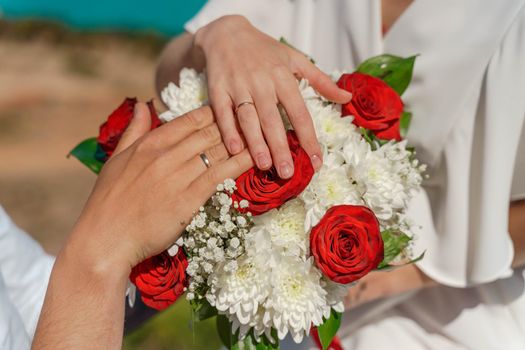 Hands of the groom and the bride with wedding rings on top of the bride's bouquet