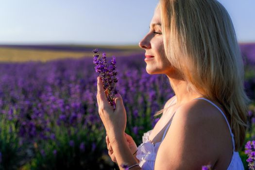 Beautiful blonde is in the field of lavender, holds a bouquet of flowers and enjoys aromatherapy. The girl's eyes are closed. The concept of aromatherapy, lavender oil, photo shoot in lavender