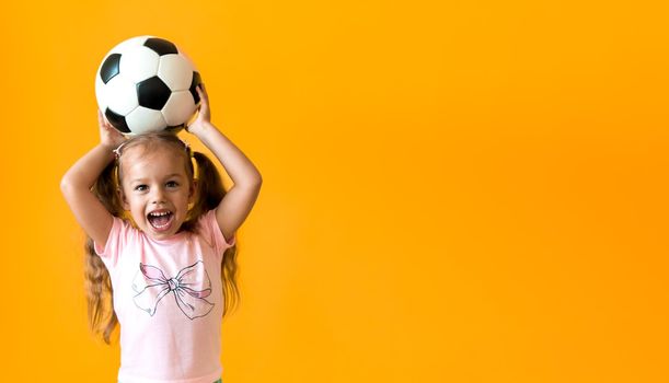 Authentic cute smiling preschool little girl with classic black and white soccer ball look at camera on yellow background. child play football in t-shirt and shorts. Sport, championship, team concept.