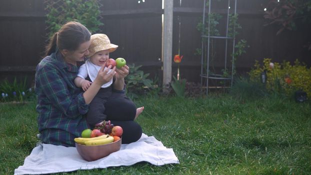 Happy Young Cheerful Mother Holding Baby Eating Fruits On Green Grass. Mom Adorable Infant Child Playing Outdoors With Love In Backyard Garden. Little Kid With Parents. Family, Nature, Ecology Concept.