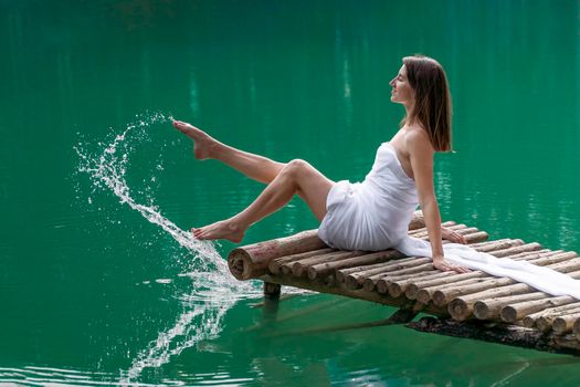 A young woman in a red light dress barefoot in the summer in a forest on a lake sits on a wooden bridge and sprays water with her foot. The concept of harmony with nature, leisure and travel, blogger