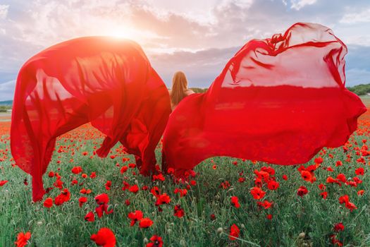 A young woman in a red dress and long red wings poses in a large field of red poppies at sunset.