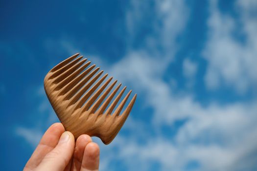 A hand holding natural wooden comb made of pear tree for scalp massage, aroma combing at blue sky background.